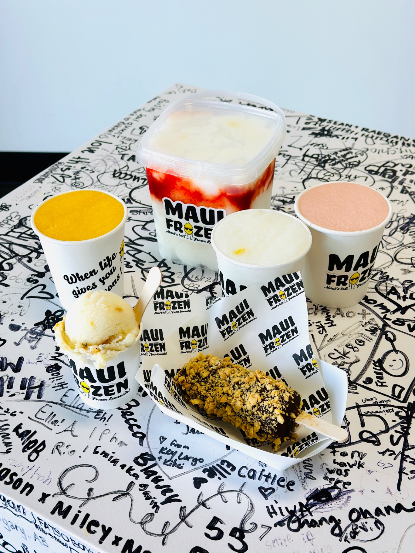 Maui Frozen product lineup: Maui Slush cups, Pineapple Scoop Ice, a pint container, and frozen bananas on the signed shop table in Kīhei.